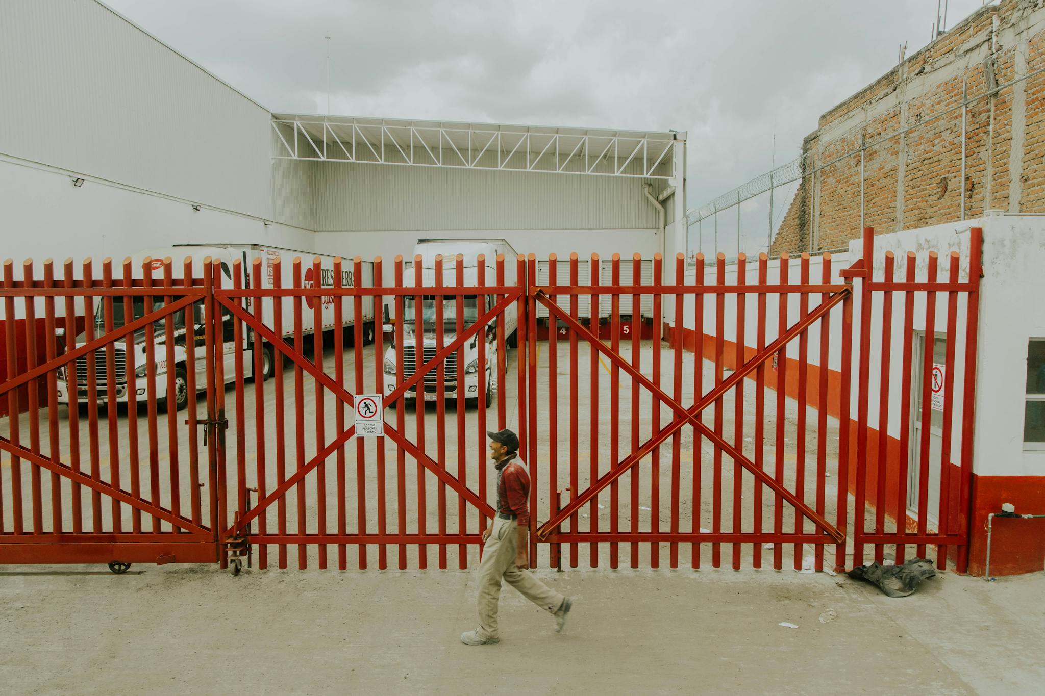 A man walks past a red industrial gate with parked trucks visible inside a facility.